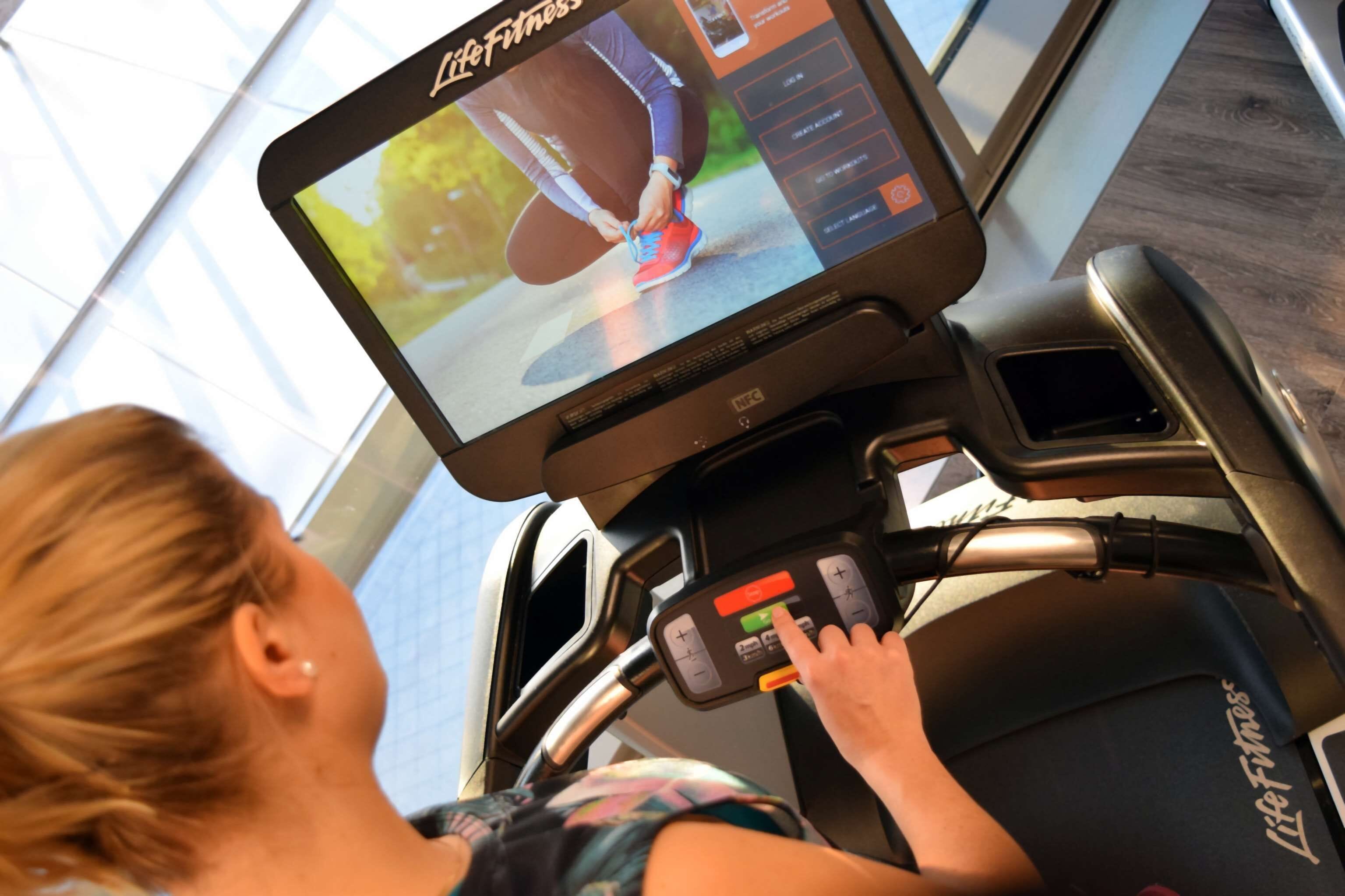 Una cinta de correr con pantalla digital que muestra a una persona corriendo, panel de control y el nombre de la marca 'Life Fitness'.