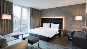 A modern hotel room with a large bed, a sofa, a small table, and a view of the city through large windows. - Hyatt Place Frankfurt Airport (Frankfurt)