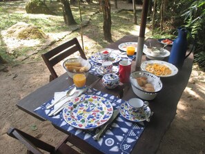 Mesa al aire libre con platos, tazones y una jarrón, rodeada de árboles.