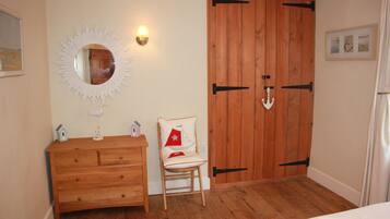 A bedroom with a wooden dresser, a chair with a red cushion, a wooden wardrobe, a wall-mounted mirror, and a small lamp.