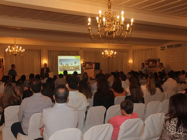 Un salón de conferencias con asistentes mirando una pantalla de presentación, candelabros y un escenario con oradores.