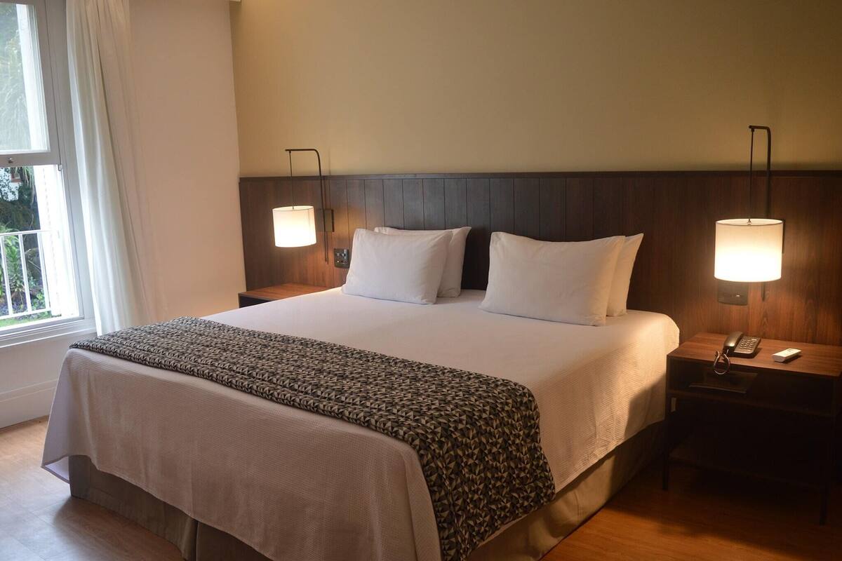 Une chambre d’hôtel avec un grand lit, deux lampes de chevet, une table de chevet avec un téléphone et une télécommande, et une fenêtre avec des rideaux.
