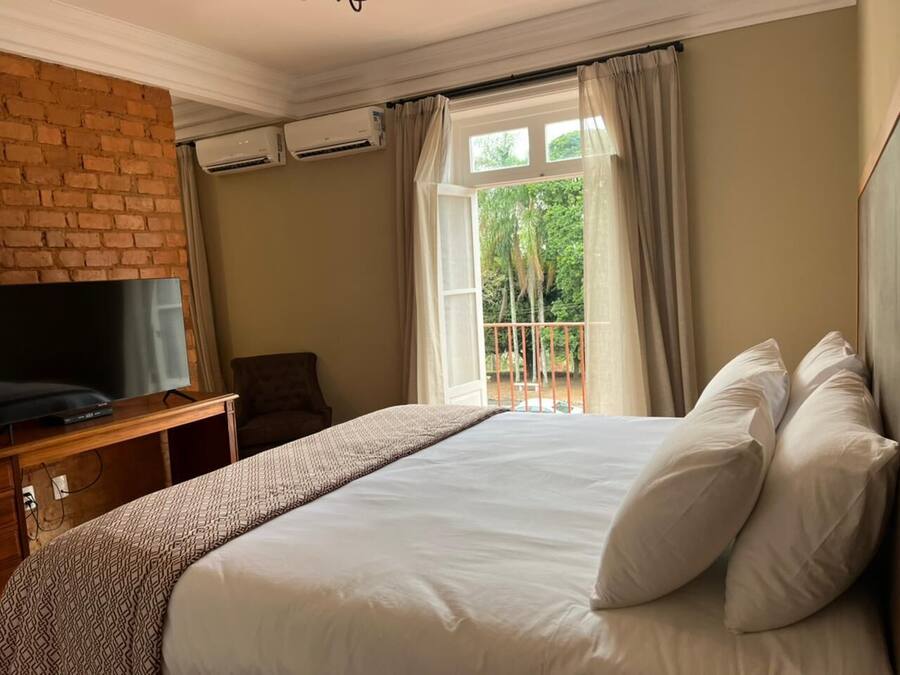 Une chambre d’hôtel avec un grand lit, un téléviseur posé sur un meuble en bois, un mur en briques et un balcon avec des rideaux.