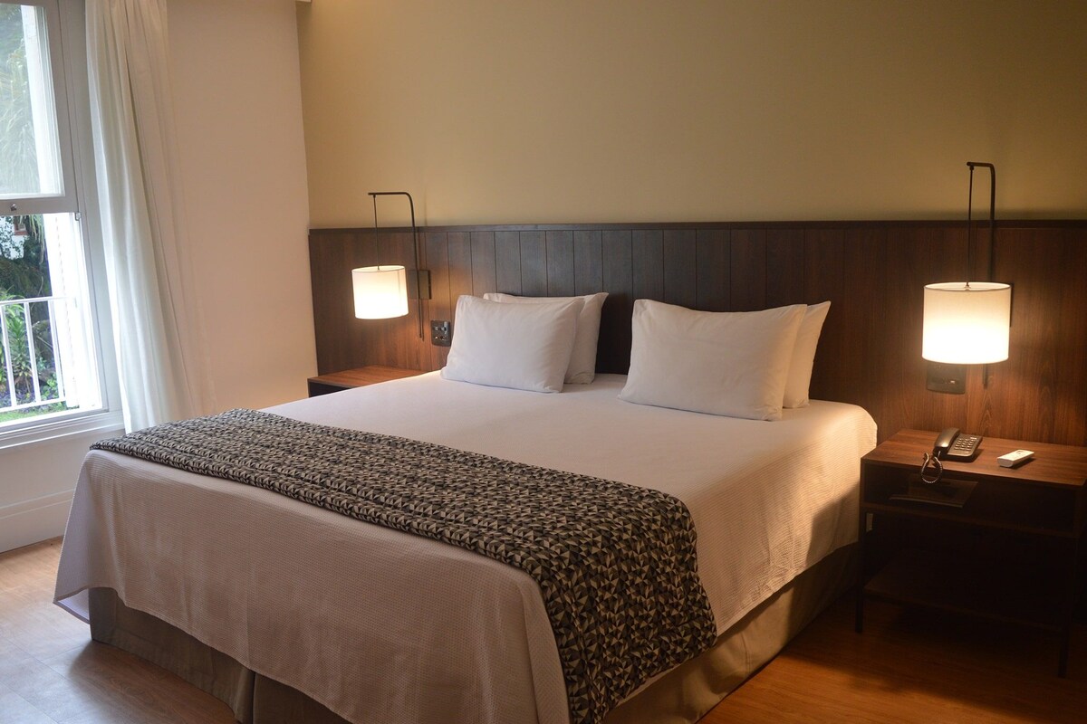 A hotel room with a large bed, two bedside lamps, a nightstand with a phone and remote, and a window with curtains.
