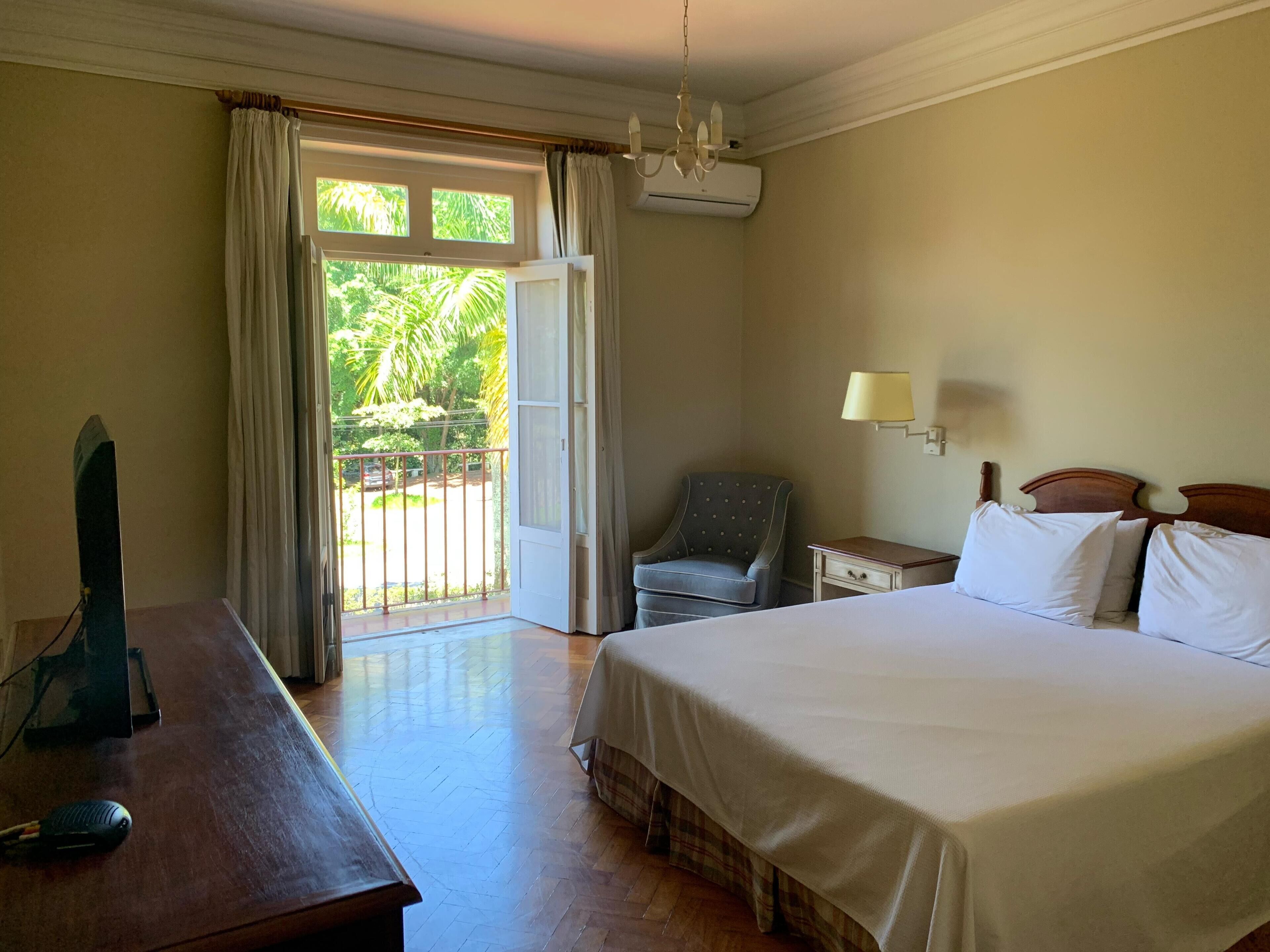Quarto de hotel com uma cama, uma escrivaninha, uma cadeira, um abajur e vista para o verde pela janela.