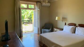 Quarto de hotel com uma cama, uma escrivaninha, uma cadeira, um abajur e vista para o verde pela janela.