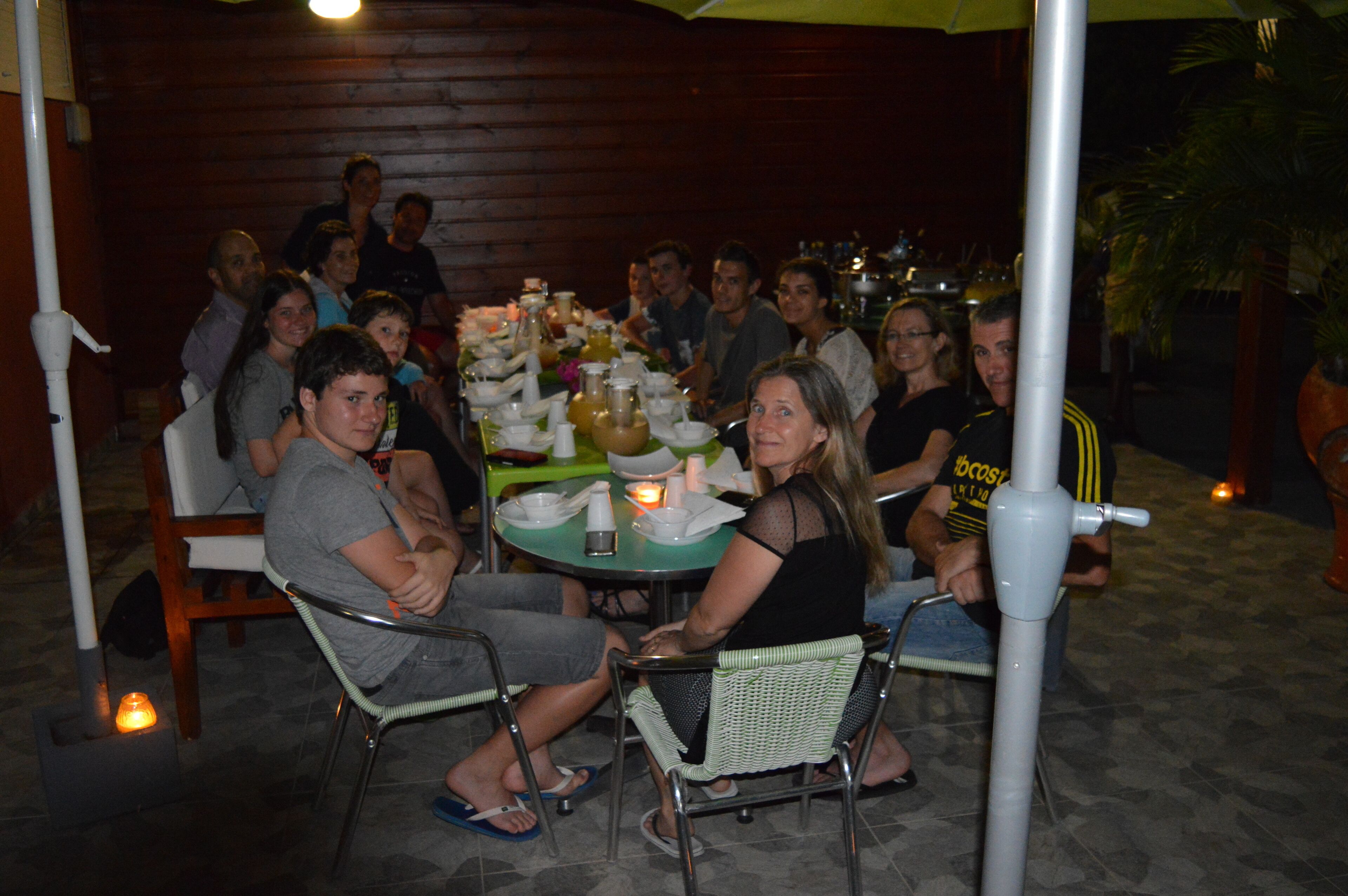 A group of people seated around a table with various dishes and drinks, in an outdoor setting with wooden walls and a tiled floor.