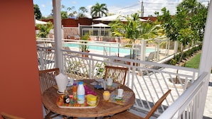 A round wooden table with chairs, a white railing, and a view of a swimming pool and palm trees. - Etal Paradise, a Tropical Paradise in Sainte-Anne in Guadeloupe (SAINTE  ANNE)