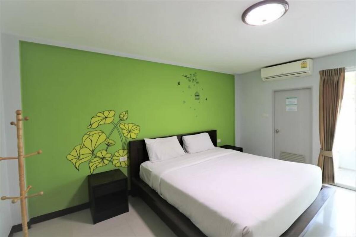 A bedroom with a green wall, a bed with white bedding, a nightstand, a wall-mounted air conditioner, and a door with a curtain.