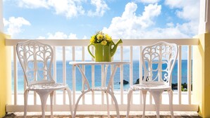 Uma varanda com cadeiras e mesa brancas, um vaso verde com flores e vista para o mar.