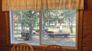 A wooden cabin interior with a dining table and chairs, a window with a view of a forest and outdoor equipment. - Gateway To Yellowstone Park And Outdoor Adventure. FREE internet and cleaning. (Island Park)