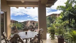 A covered patio with a wooden table and chairs, surrounded by potted plants and a view of a large building in the background. - Luxury Los Sueños Condo - Marina, Pools & Golf. Fishermans Paradise! (Los Suenos)