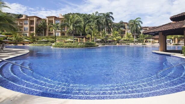 A large, clear swimming pool surrounded by palm trees and a resort building. - Luxury Los Sueños Condo - Marina, Pools & Golf. Fishermans Paradise! (Los Suenos)