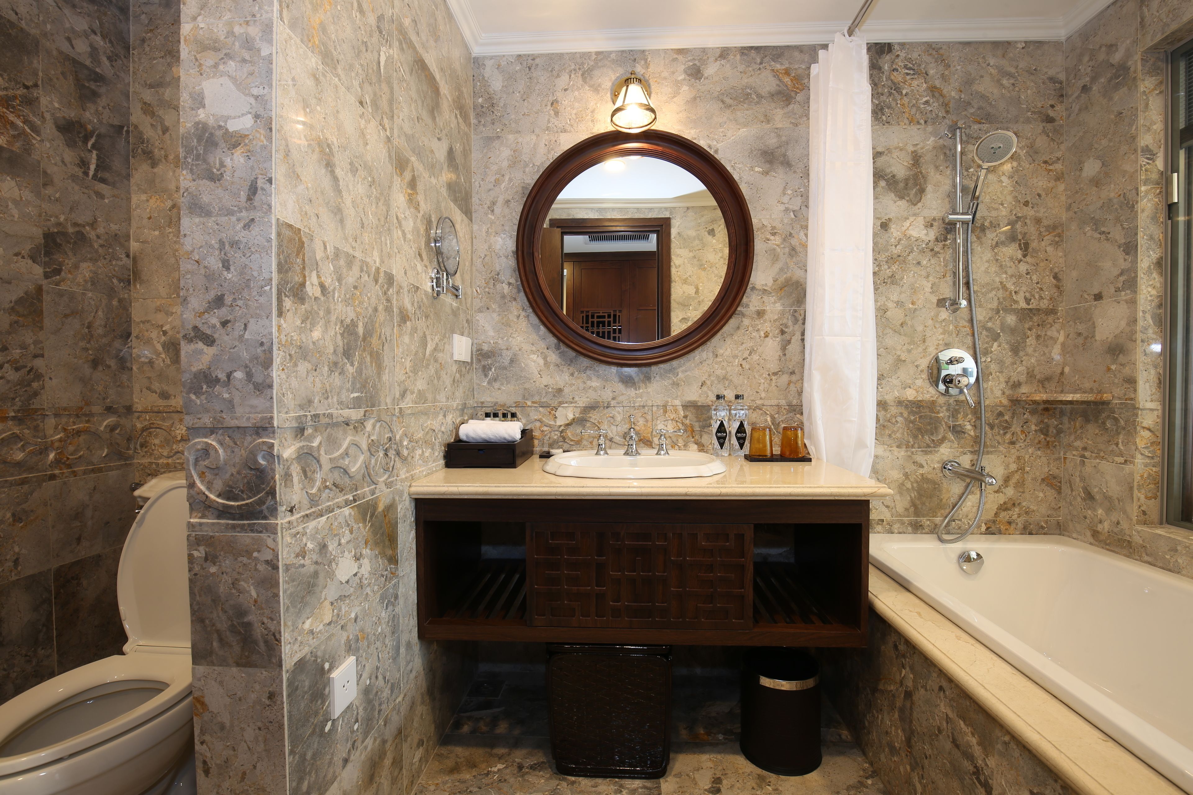 Un baño con pared de mármol, un espejo redondo, un lavamanos, una bañera y un inodoro.