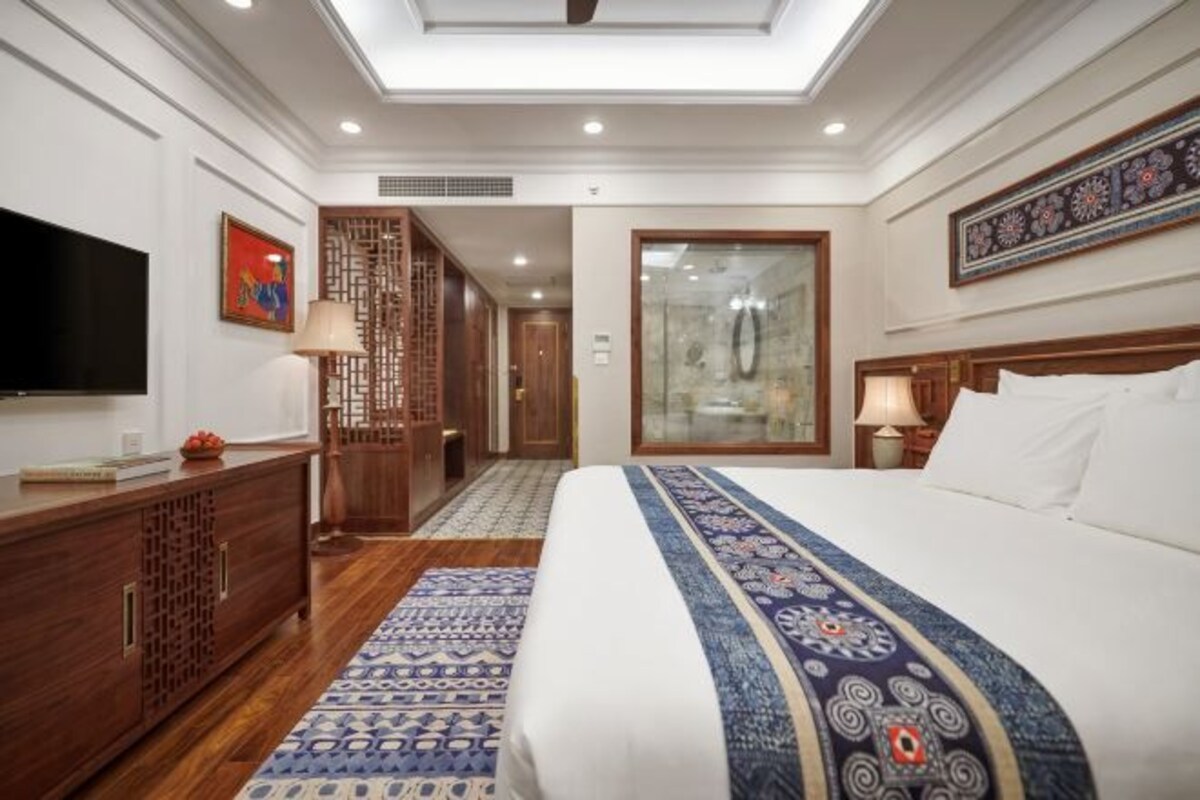 A hotel room with a large bed, a television, a wooden cabinet, a patterned rug, and a decorative wall piece.