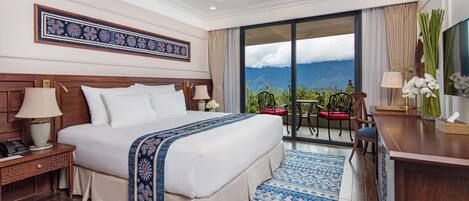 A hotel room with a large bed, a desk, a chair, a TV, and a balcony with a mountain view.