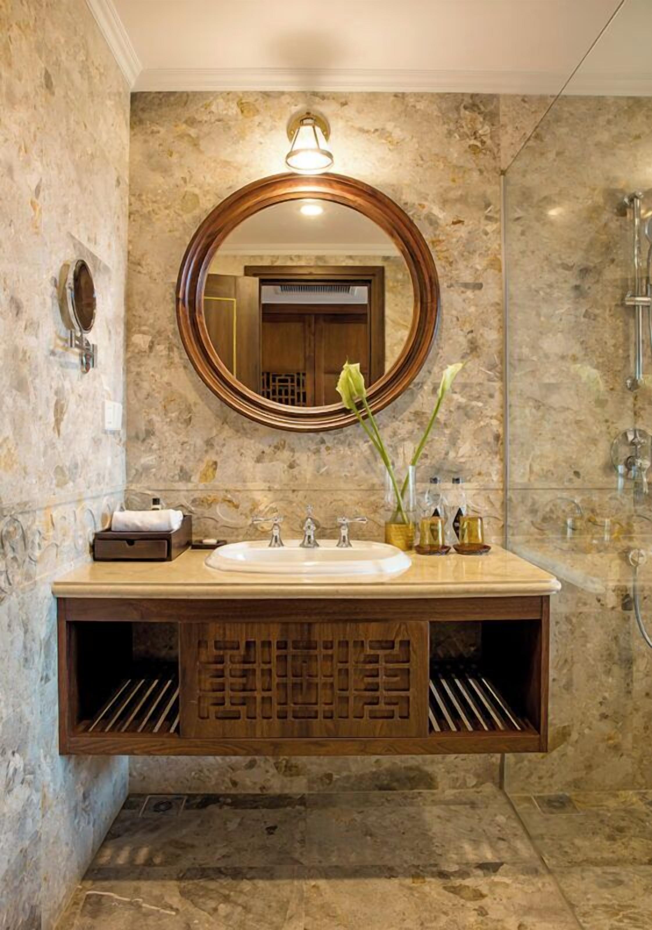 A bathroom with a marble wall, a wooden vanity, a round mirror, and a shower area.