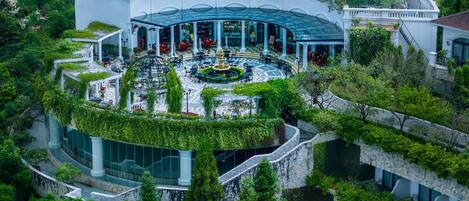 Un edificio grande y blanco con un patio central, rodeado de abundante vegetación y muros de piedra.