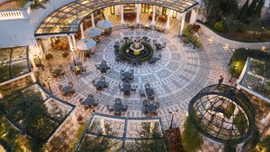 Une vaste cour extérieure dotée d’une fontaine centrale, entourée de plusieurs espaces de détente et surmontée d’un niveau supérieur entièrement vitré.