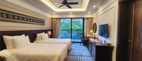 A hotel room with two beds, a desk with a chair, a television, and a balcony with a view of greenery.