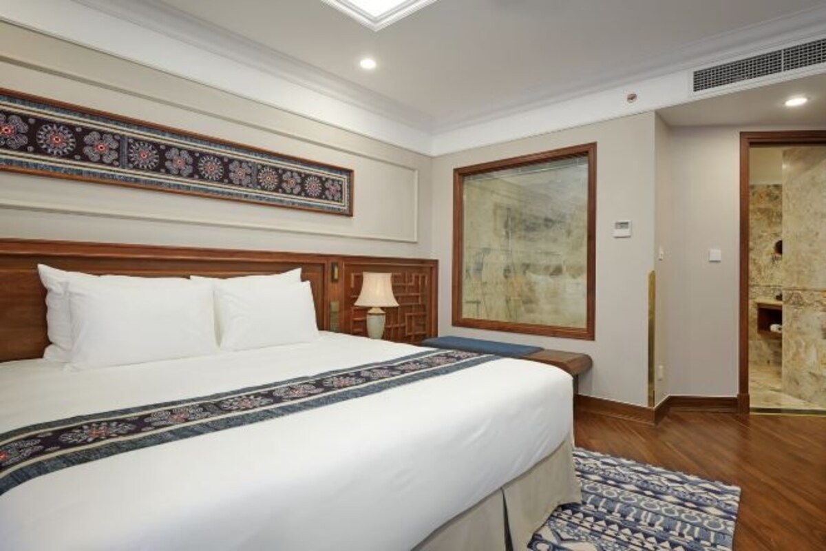 A hotel room with a large bed, a wooden headboard, a decorative wall panel, a large window, and a bathroom with a stone wall.