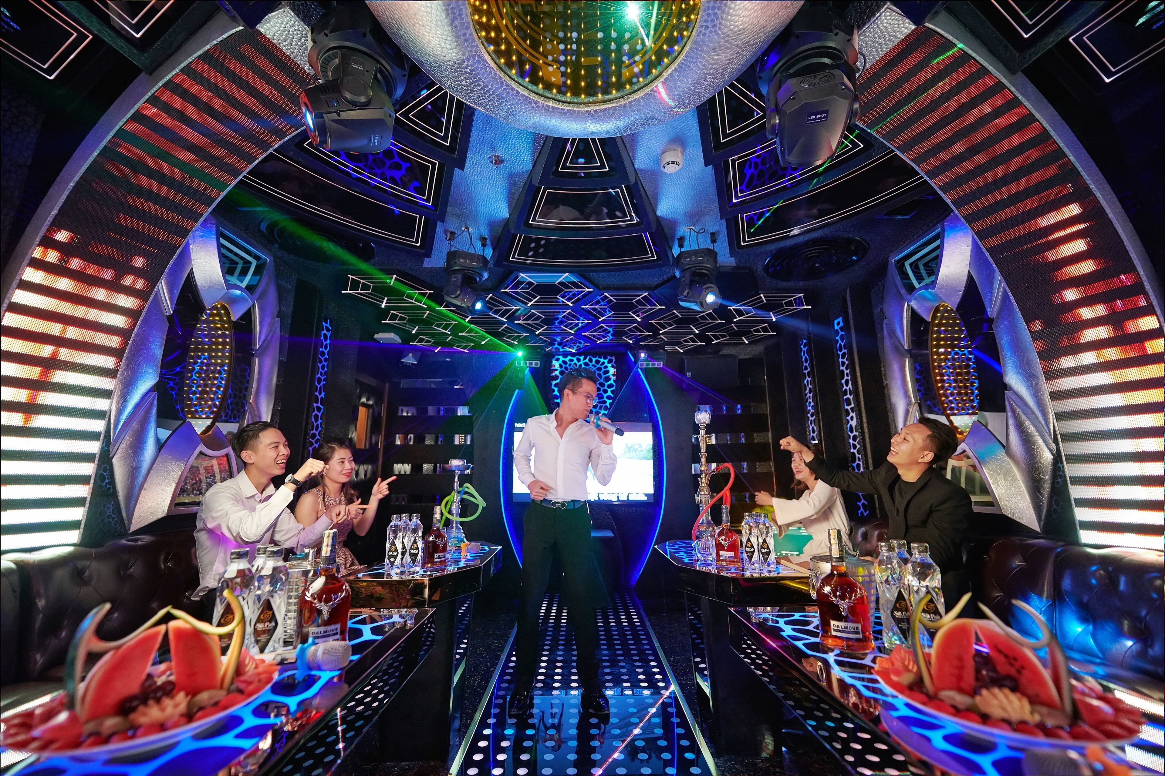 Een helder verlichte, moderne bar met een centraal podium, tafels met drankjes en hapjes, en een geometrisch plafondontwerp.