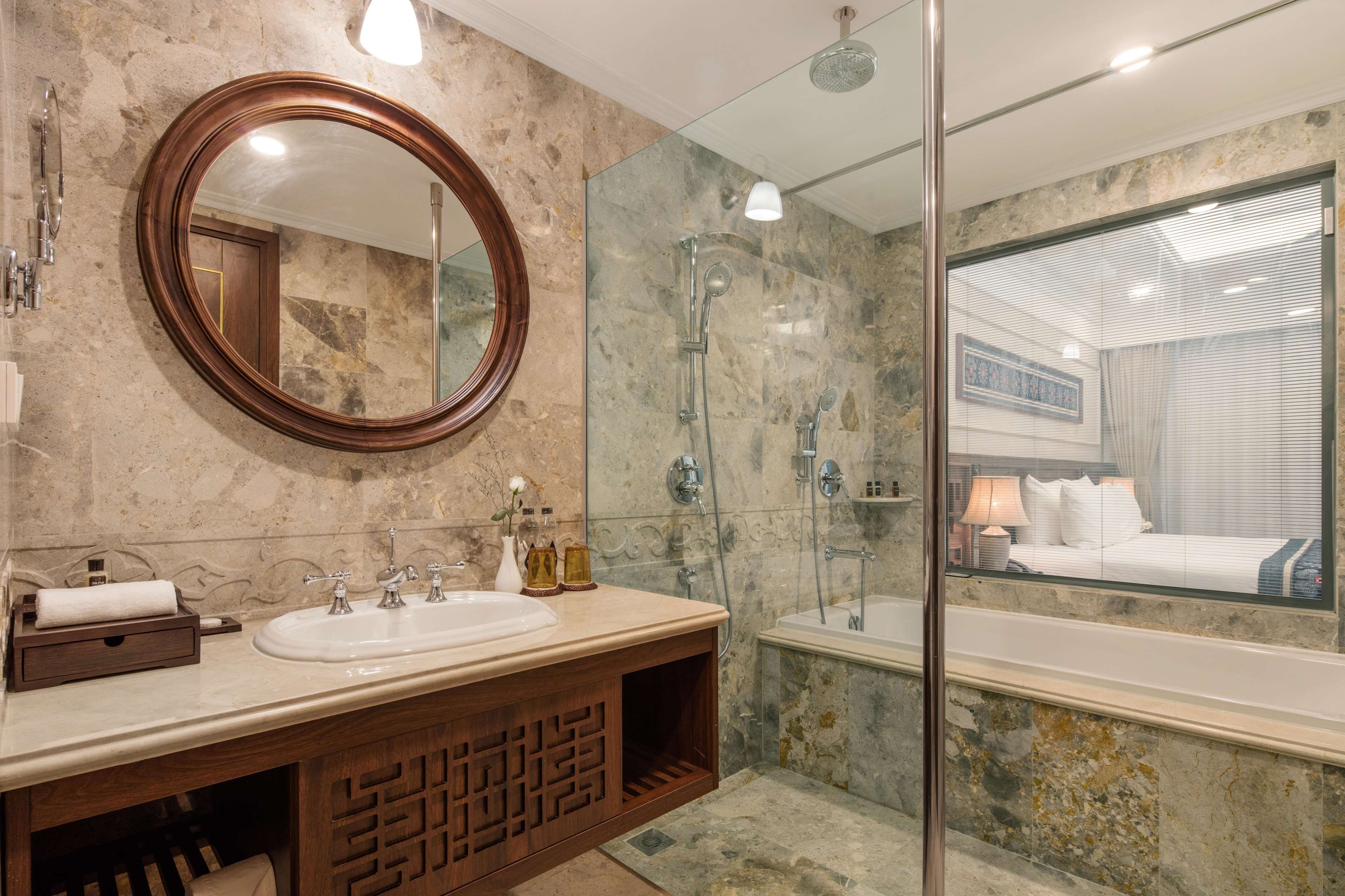 Un baño con lavamanos de mármol, tocador de madera, un espejo grande, cabina de ducha de vidrio y vistas a un dormitorio a través de un panel de vidrio.