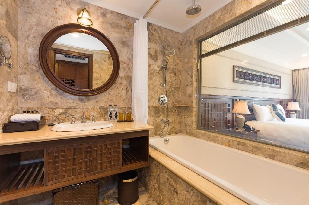 Un baño con pared de m ármol, espejo redondo, lavamanos, ducha y un ventanal grande que permite ver una cama.