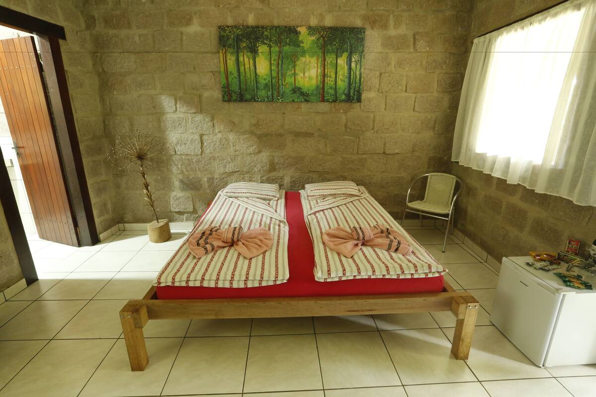 Una habitación con una cama, una mesita, una silla, una planta en maceta y un cuadro en la pared.