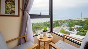 A room with a large window offering a view of a cityscape, a framed poster on the wall, and a small table with two cups and a potted plant. - GAIA Hotel & Residence Phu Quoc (Phu Quoc)