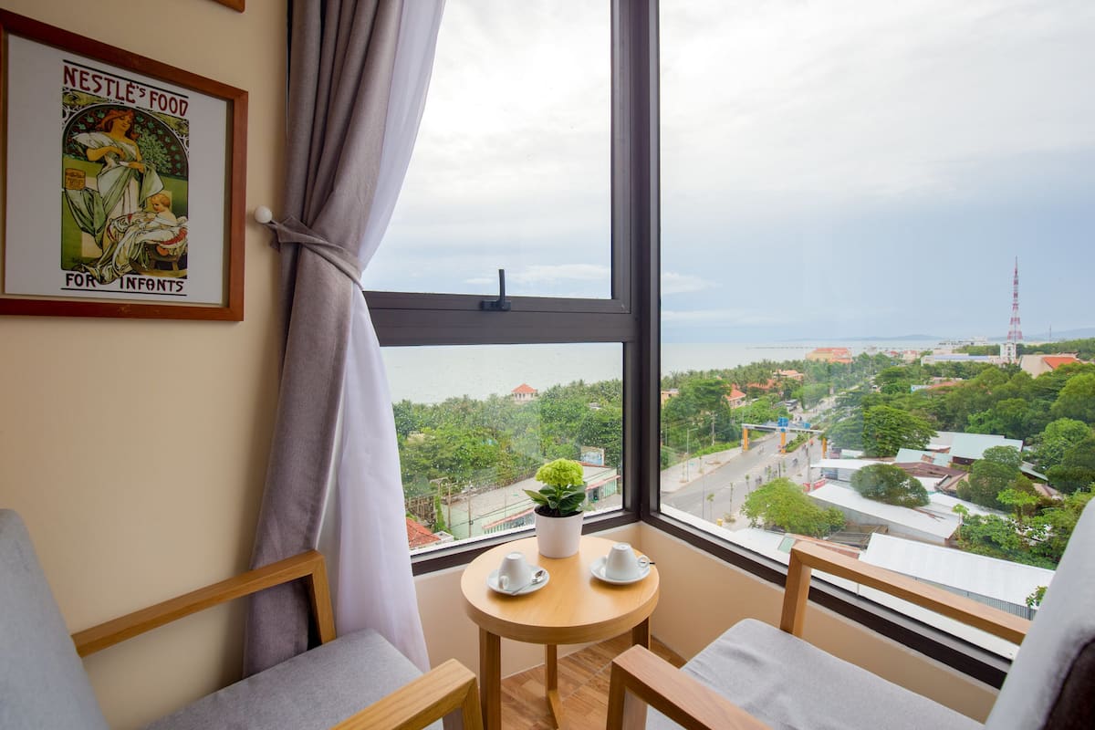 A room with a large window offering a view of a cityscape, a framed poster on the wall, and a small table with two cups and a potted plant.
