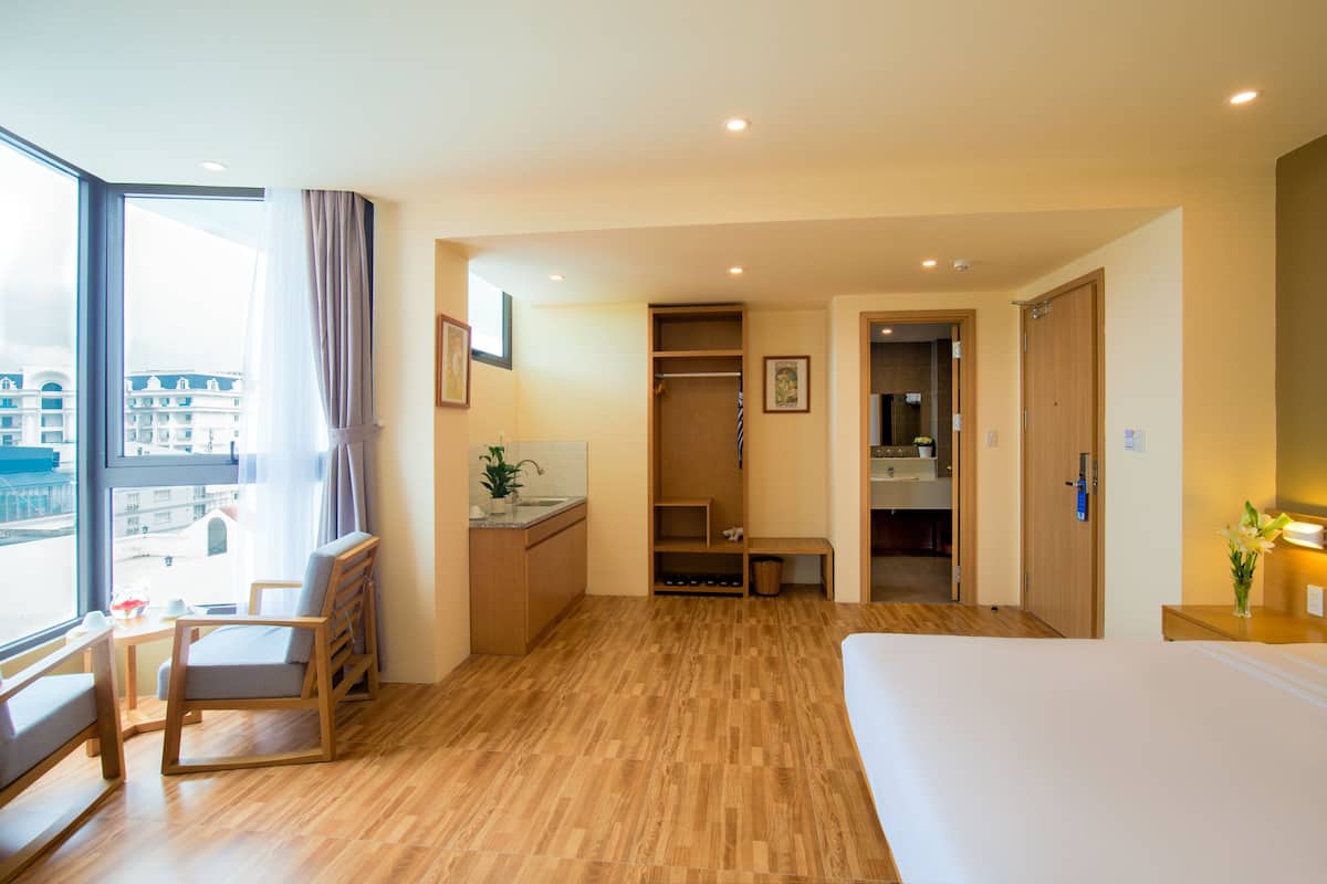 A hotel room with a large bed, wooden flooring, a desk with a chair, a closet, and a view of the city through a large window.