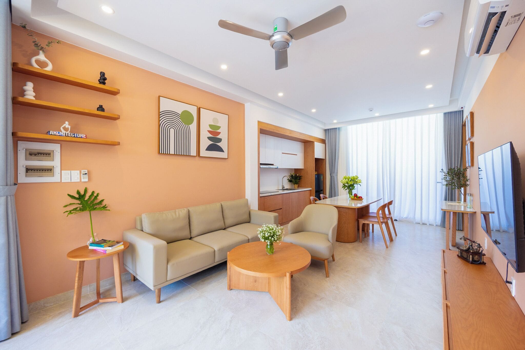 Una sala moderna con sofá, mesa de centro y comedor, que incluye un ventilador de techo, estantes montados en la pared y una cocina al fondo.