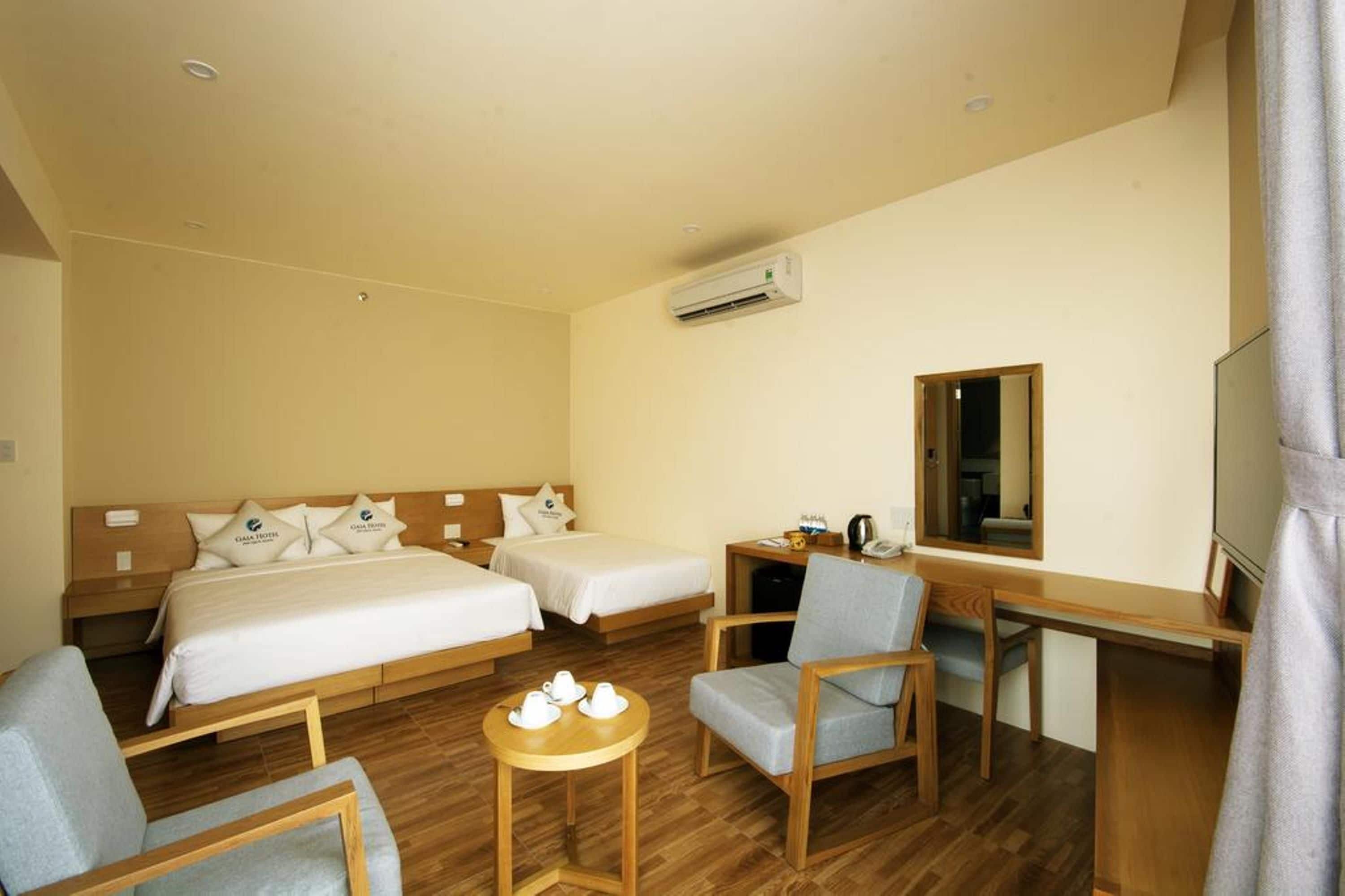 A hotel room with two beds, a desk, a chair, a small table with cups, and an air conditioning unit.