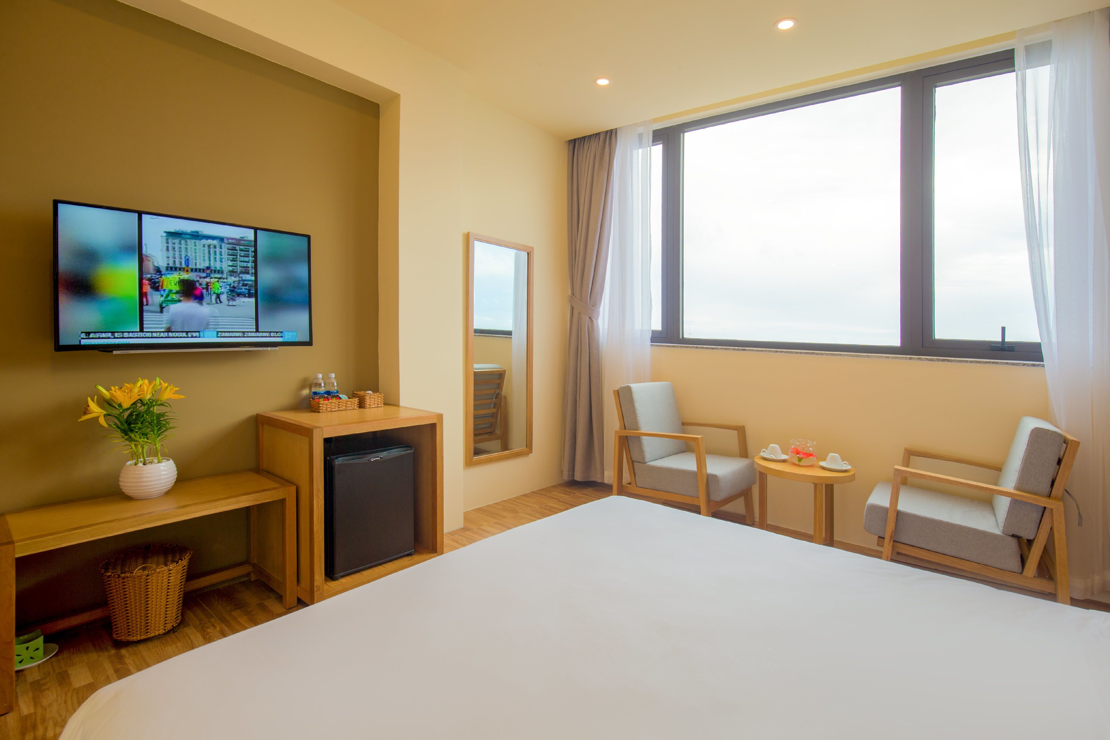 A hotel room with a large bed, a television mounted on the wall, a small desk with a refrigerator, a chair, and a window with curtains.