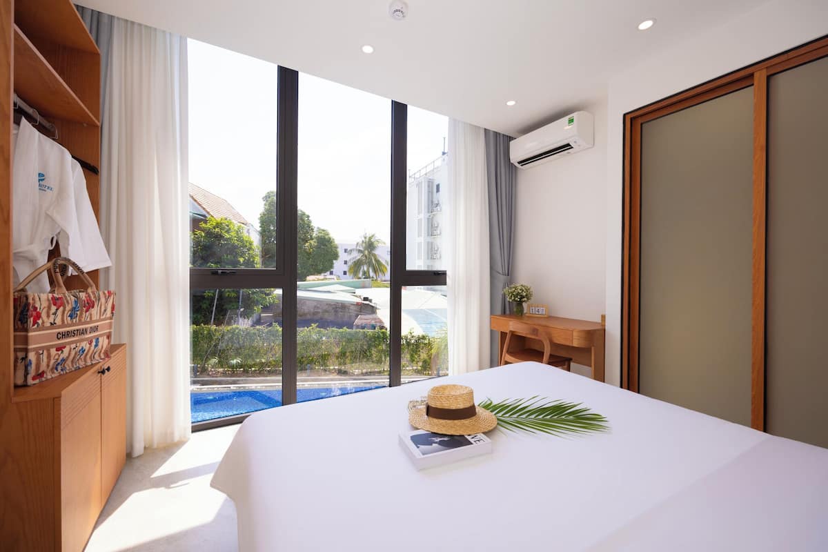 A hotel room with a large window, a bed, a wooden wardrobe, and a small table.