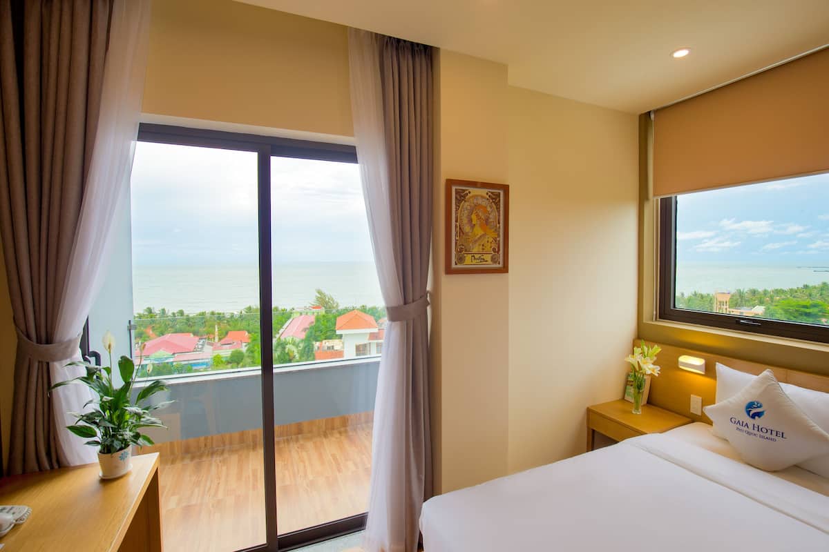 A hotel room with a bed, a nightstand, a window with curtains, a view of the sea, and a painting on the wall.