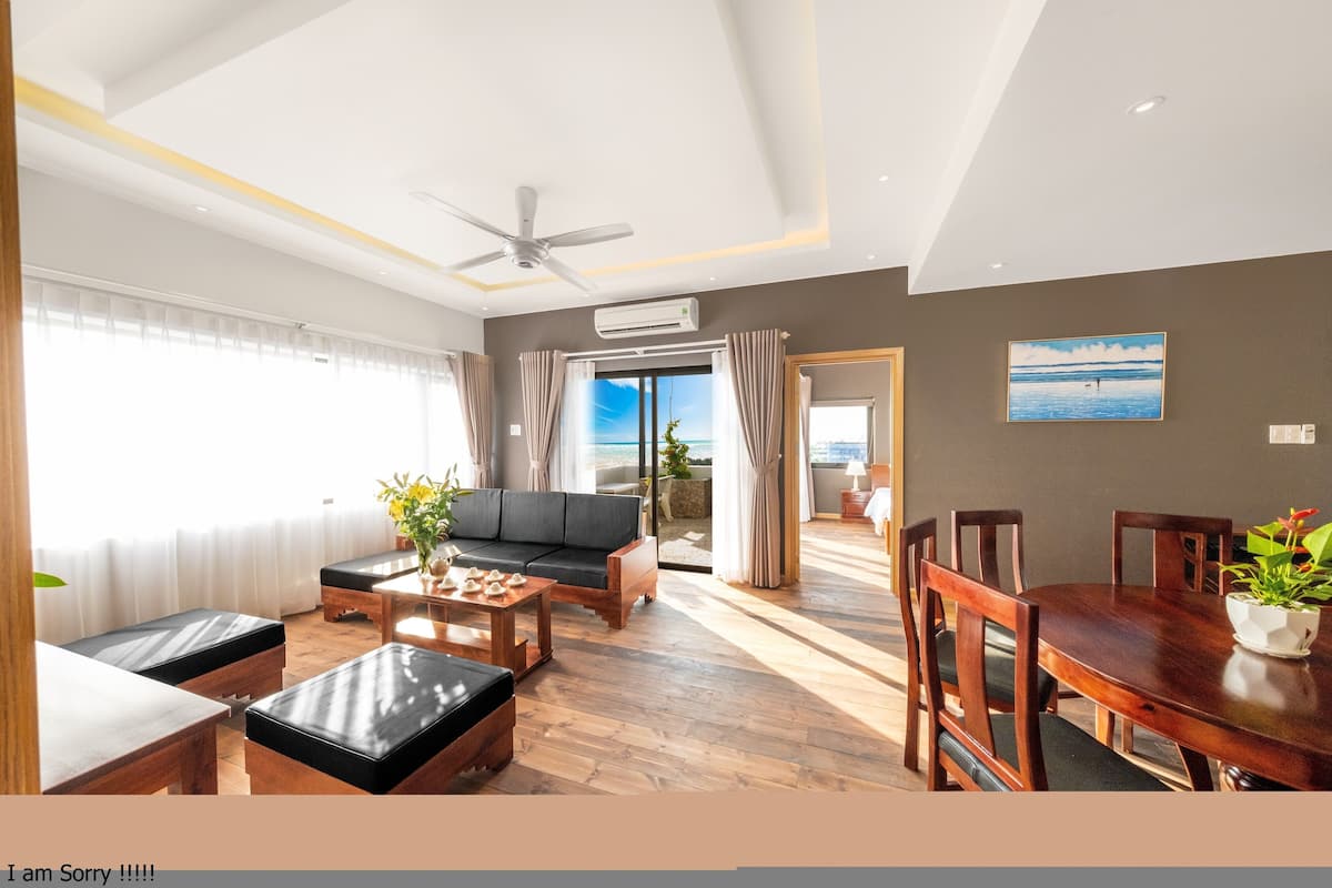 A spacious living room with a sofa, coffee table, and dining table. There is a large window with curtains, a ceiling fan, and an air conditioning unit.