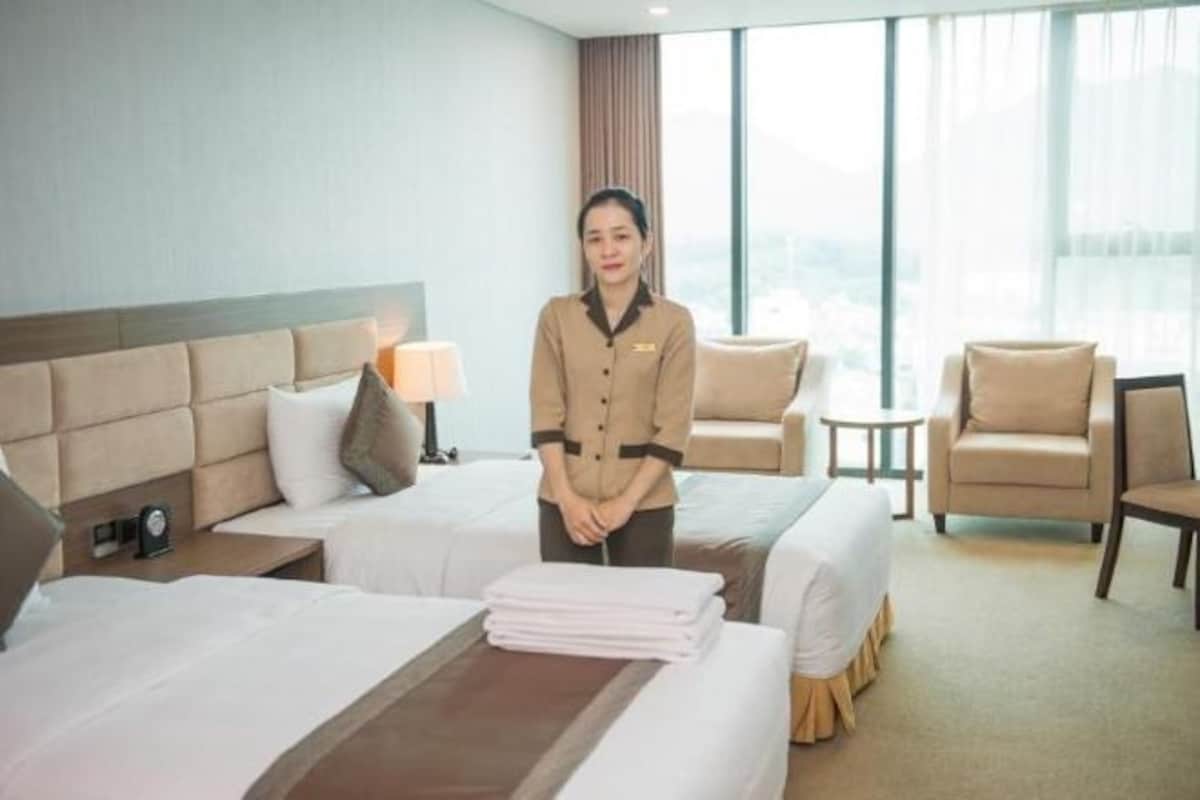 Habitación de hotel con dos camas, una mesita de noche con lámpara, una silla y vistas a la ciudad a través de amplias ventanas.