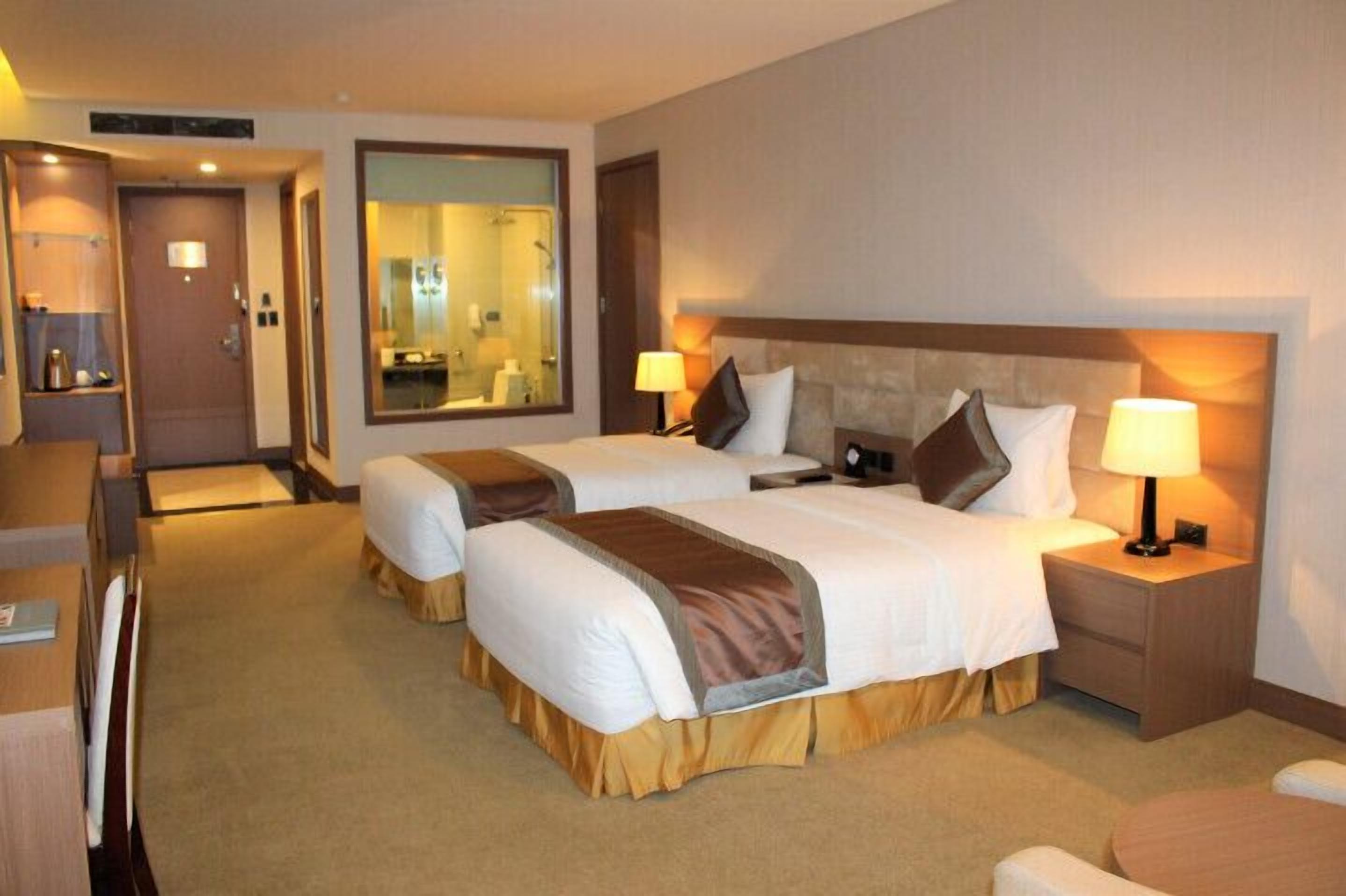 Une chambre d’hôtel avec deux lits, un bureau, un miroir et une salle de bain visible à travers une porte ouverte.