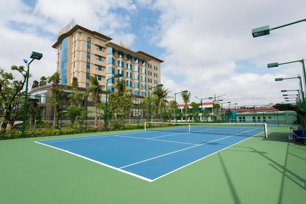 A tennis court with a net, surrounded by green fencing and palm trees.