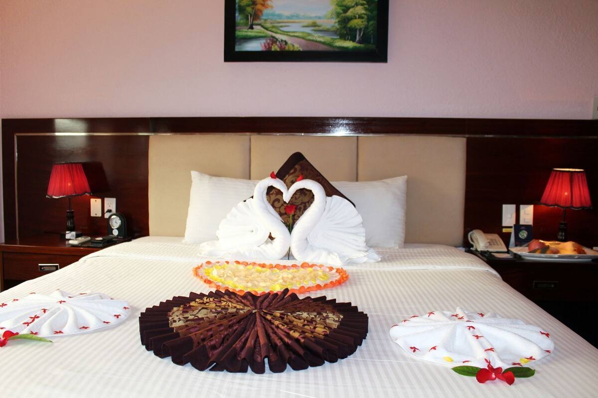 A hotel room with a neatly made bed, a decorative swan centerpiece, and a painting on the wall.
