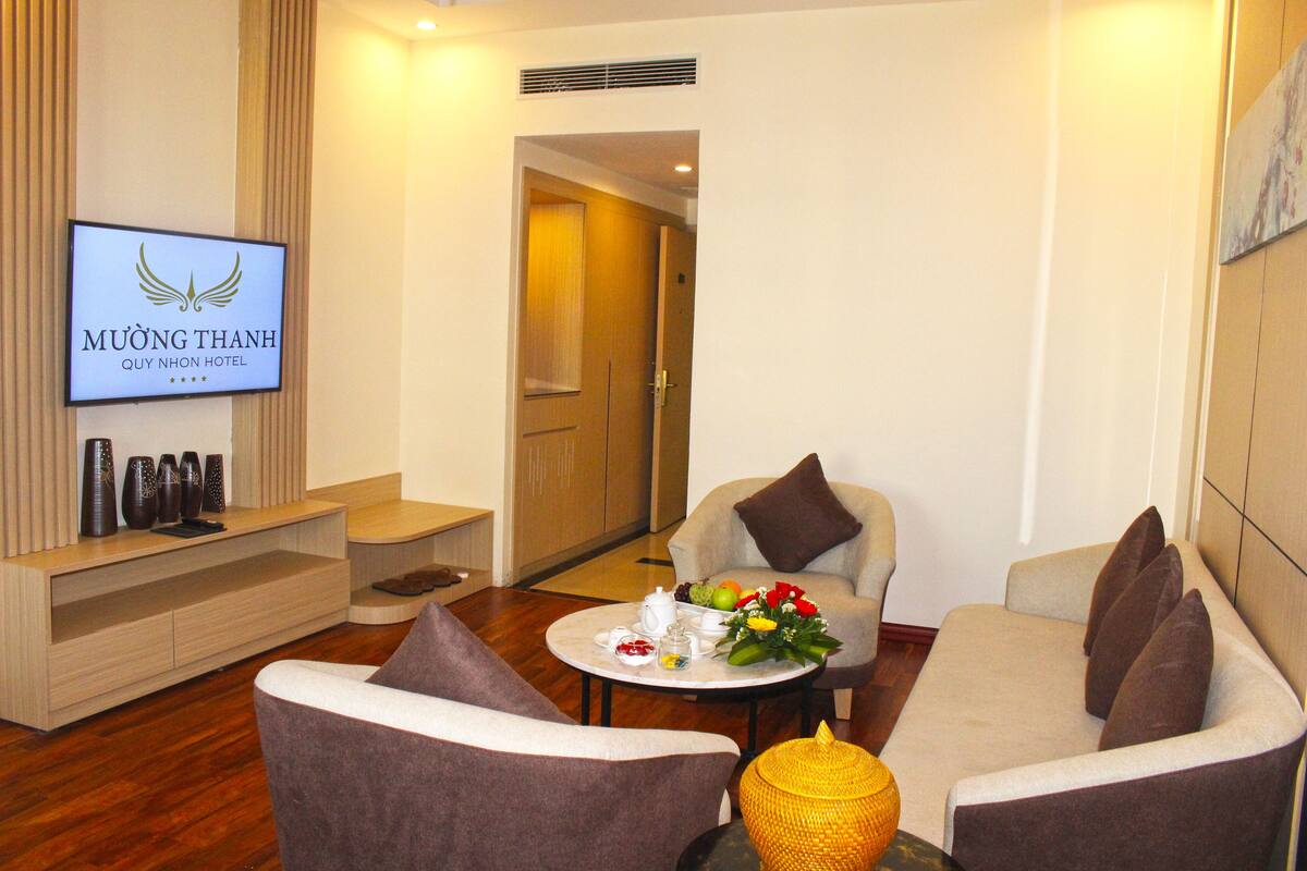 A modern hotel lobby with a flat-screen TV displaying the hotel name, comfortable seating, a small table with a floral arrangement, and a decorative vase.