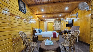 A cozy cabin-style room with a wooden interior, a bed with white linens, two wicker chairs, a small table with a bottle and a cup, and a television on the wall. - Ankit Vista Green Village (Nelamangala)