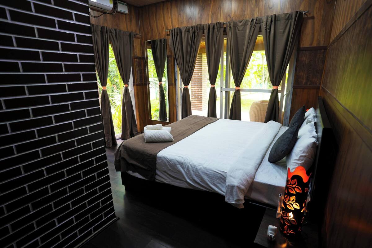 A bedroom with a brick wall, a bed with white and brown bedding, and curtains on the windows.
