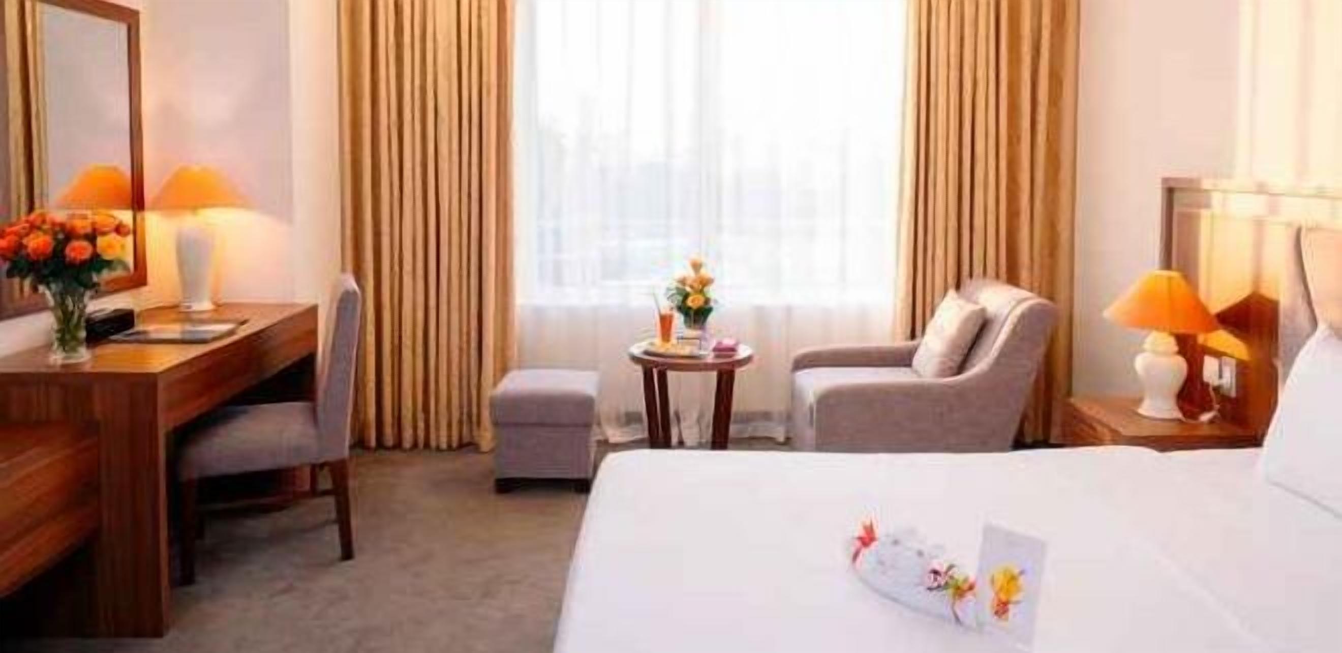 A hotel room with a bed, a desk, a chair, a small table, and a lamp.