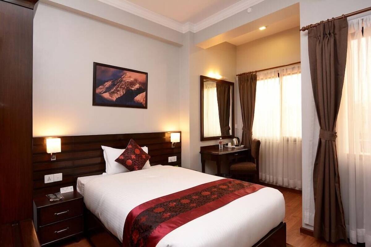 A hotel room with a bed, bedside lamps, a desk, a chair, a mirror, and a window with curtains.
