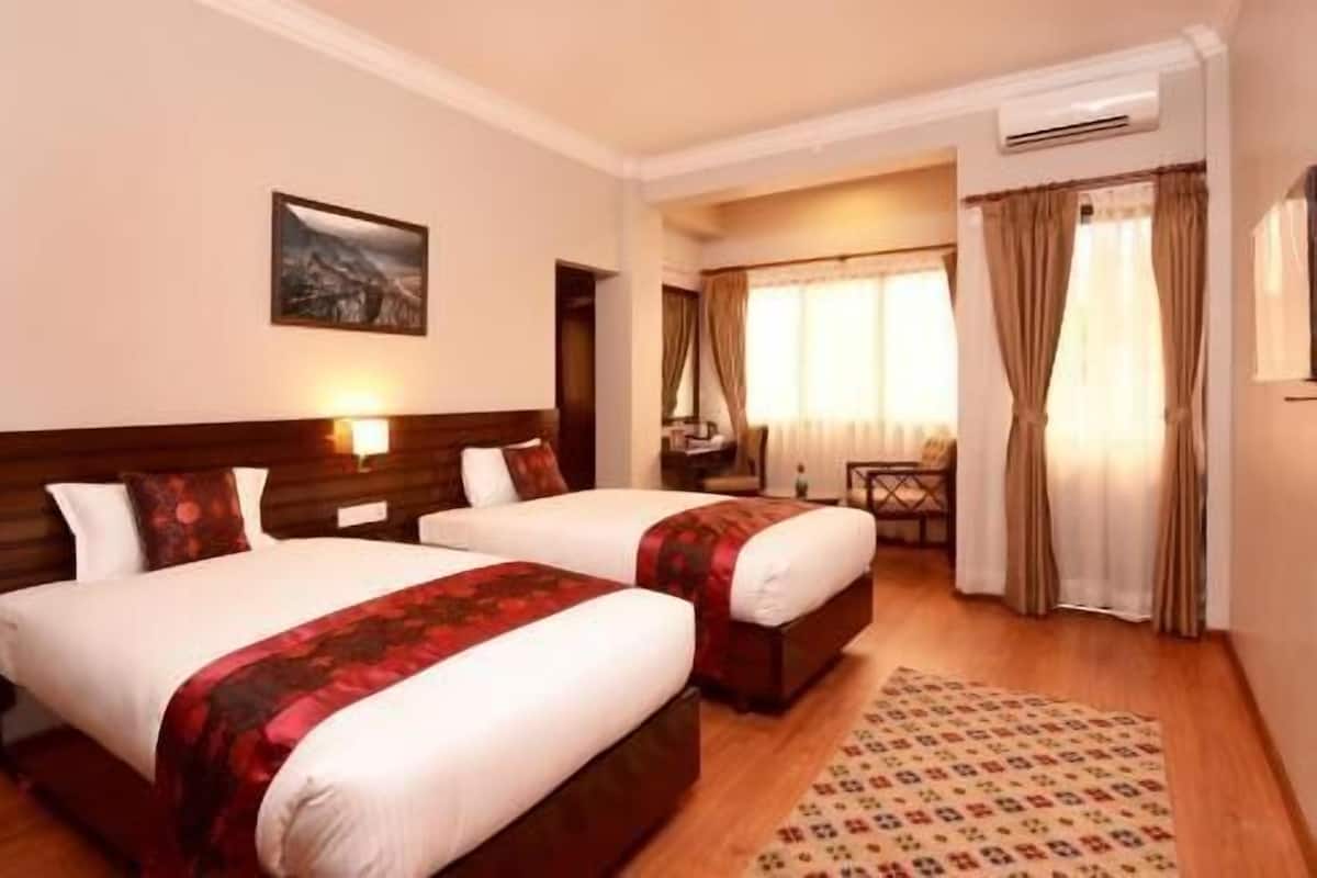 A hotel room with two beds, a desk, a chair, a painting on the wall, and an air conditioning unit.