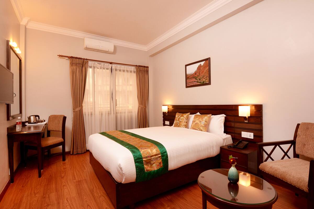 A hotel room with a large bed, a desk with a chair, a bedside table, a TV, and a painting on the wall.