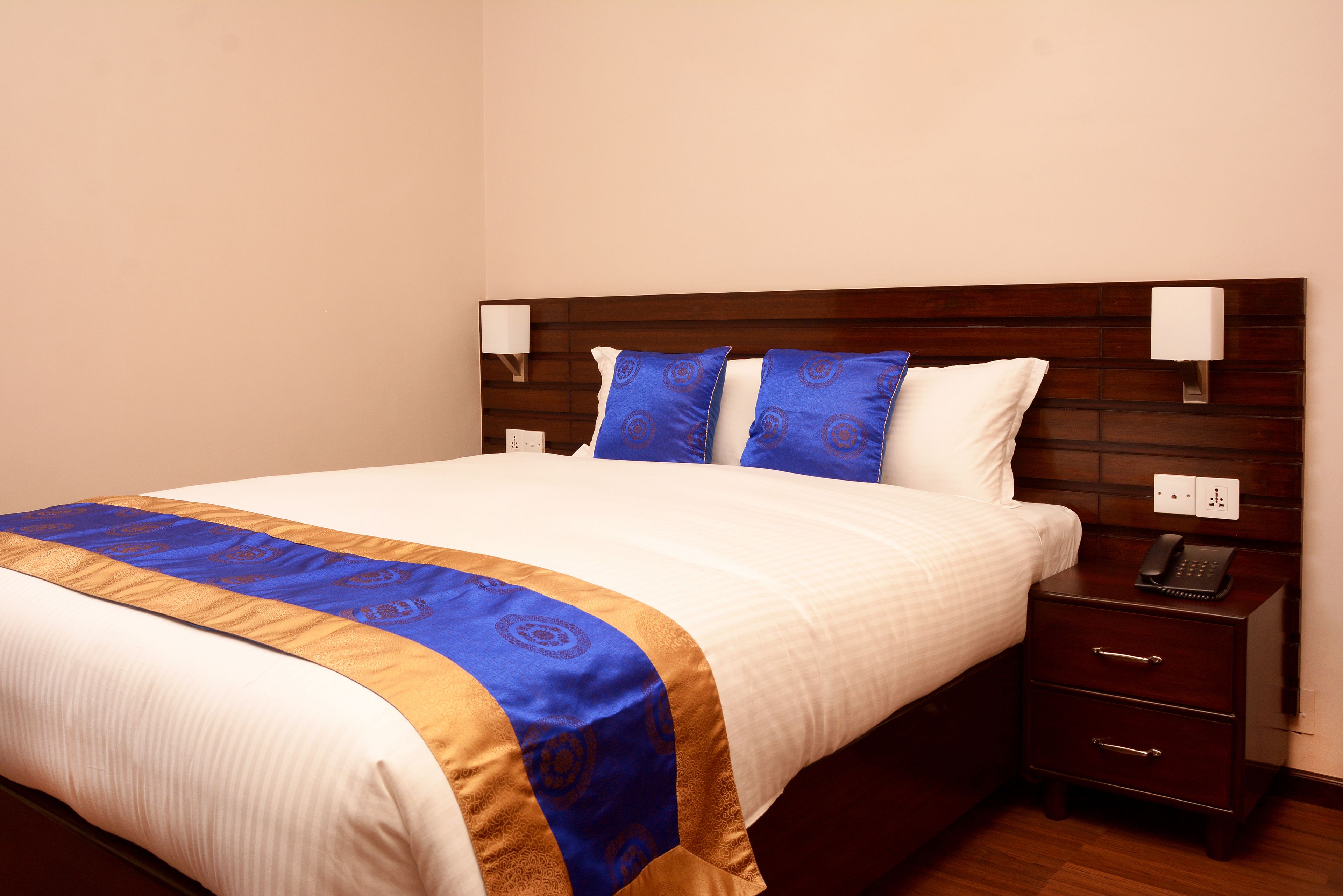 Una cama bien hecha con una colcha blanca y azul, un cabecero de madera y una mesita de noche con un teléfono y una lámpara.